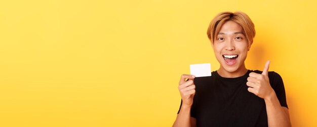 Foto close-up van een gelukkige en tevreden aziatische knappe man die zijn creditcard toont en zijn duim omhoog in goedkeuring glimlacht verbaasd staande gele achtergrond persoon