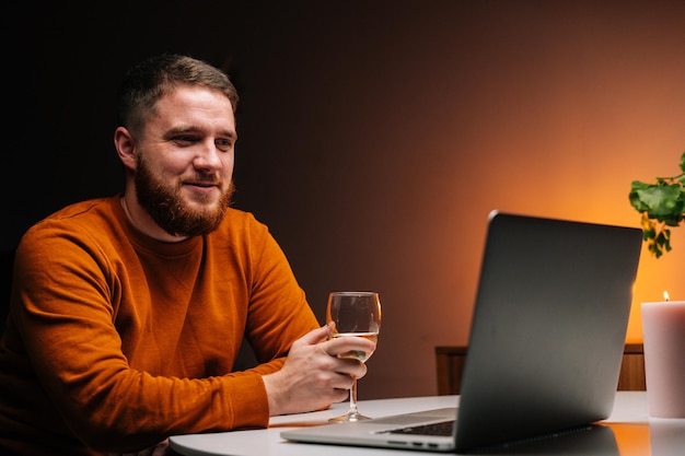 Close-up van een gelukkige jonge man die geniet van daten op afstand met behulp van een laptopcomputer, glas met champagne vasthoudt, pratend roosteren via videogesprek. Paar verliefd chatten op externe datum.