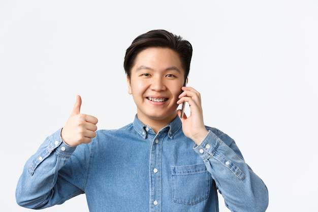 Close-up van een gelukkige tevreden aziatische man die zijn duim omhoog laat zien terwijl hij aan de telefoon praat, een positief antwoord of toestemming ontvangt, tevreden glimlacht, iets aanbeveelt, witte achtergrond.