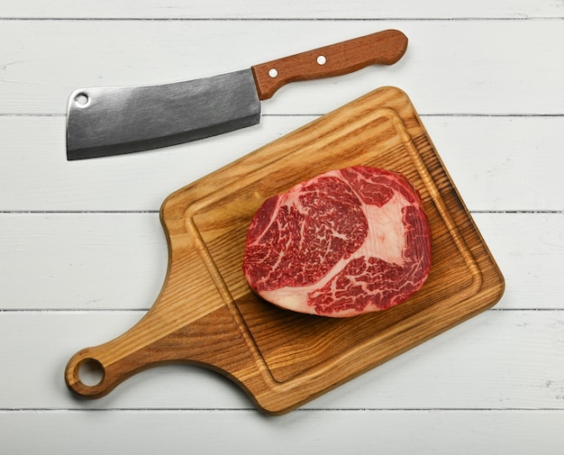 Close-up van een gemarmerde rauwe ribeye biefstuk op bruin eikenhouten snijplank met hakmes mes, over witte houten tafel, verhoogde bovenaanzicht, direct erboven