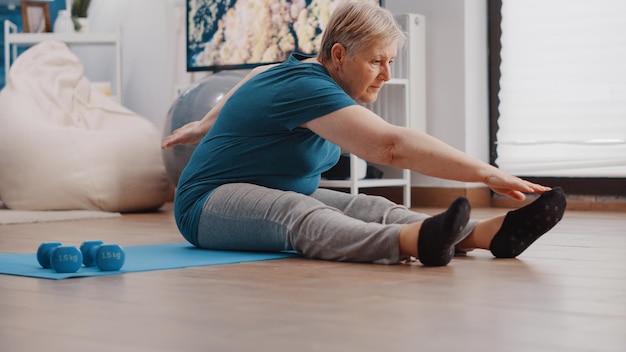 Close-up van een gepensioneerde die lichaamsbeweging doet om de spieren thuis te strekken. Gepensioneerde vrouw die armen en benen strekt terwijl ze op yogamat zit. Senior persoon training met fitness activiteit