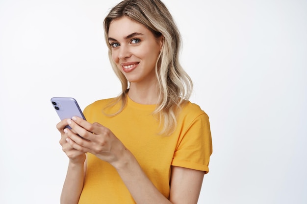 Close-up van een glimlachend mooi meisje dat een gadget voor een mobiele telefoon gebruikt en gelukkig naar de camera kijkt die in een gele t-shirt staat op een witte achtergrond