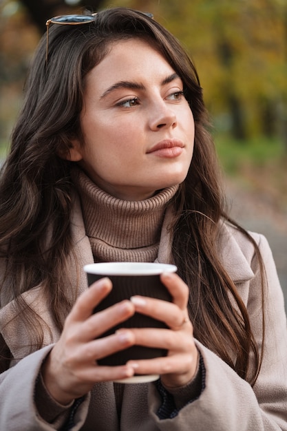 Close-up van een glimlachende jonge aantrekkelijke vrouw die een herfstjas draagt die afhaalkoffie buitenshuis drinkt