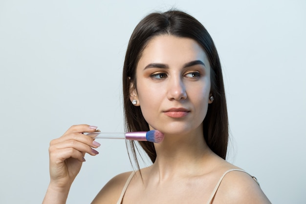 Close-up van een jong meisje in een lichte top op een witte achtergrond die een gezichtsmake-up maakt. Een mooie vrouw houdt een cosmetische borstel bij haar gezicht en glimlacht.