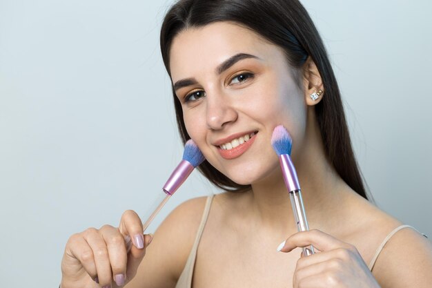 Close-up van een jong meisje in een lichte top op een witte achtergrond die een gezichtsmake-up maakt Een mooie vrouw houdt een cosmetische borstel in de buurt van haar gezicht en glimlacht