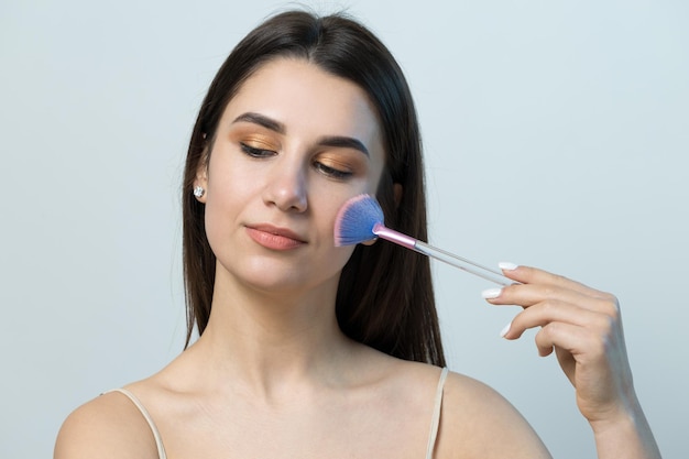 Close-up van een jong meisje in een lichte top op een witte achtergrond die een gezichtsmake-up maakt Een mooie vrouw houdt een cosmetische borstel in de buurt van haar gezicht en glimlacht