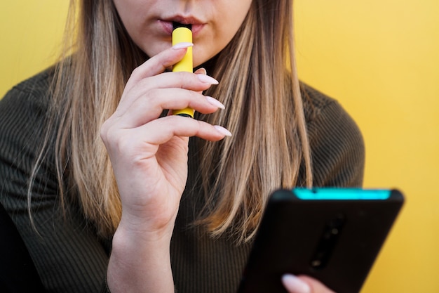 Close up van een jong meisje rookt een wegwerp elektronische sigaret. Helder gele achtergrond. Alternatief voor gewone sigaretten en vape