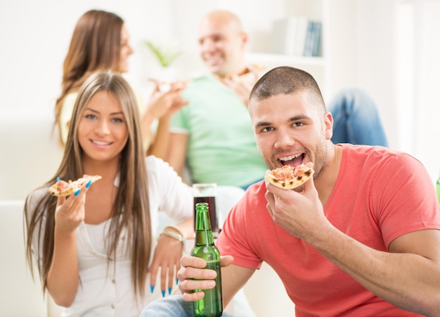 Close-up van een jonge man die lacht en pizza eet en een beer drinkt met haar vrienden op de achtergrond.