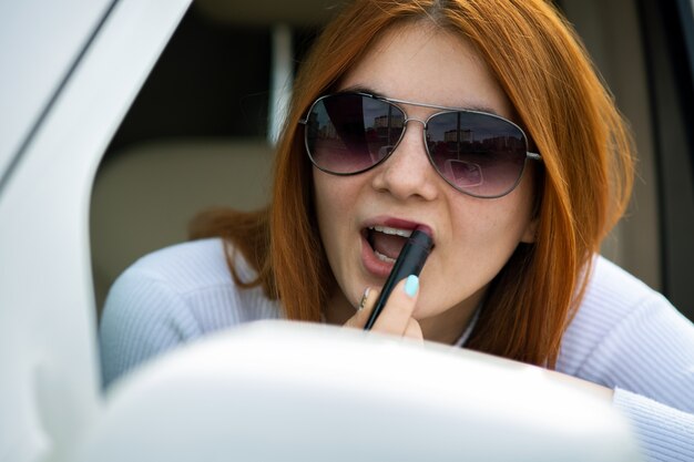 Close-up van een jonge roodharige vrouwenbestuurder die haar make-up met donkerrode lippenstift corrigeert die in autoachteruitkijkspiegel achter het stuur van een voertuig kijken.