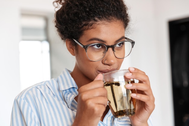 Close-up van een lachende jonge Afrikaanse zakenvrouw die een kopje thee drinkt terwijl ze thuis staat