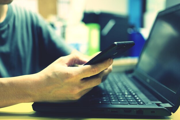 Foto close-up van een man die een mobiele telefoon gebruikt boven een laptop aan tafel