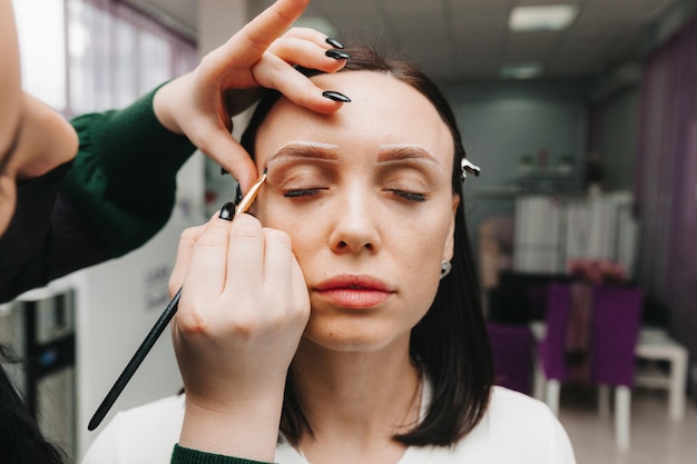 Close-up van een meisjesgezicht de meester houdt een wit potlood vast en tekent de vorm van de wenkbrauwen voor permanente make-up