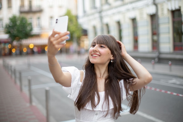 Close-up van een mooi jong meisje in witte jurk met lang bruin haar die lacht en selfie foto neemt op mobiele telefoon op de achtergrond van de straat in de zomer