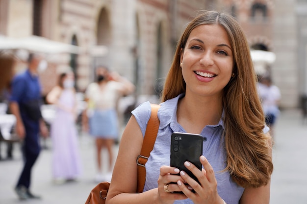 Close-up van een mooie zelfverzekerde jonge vrouw die haar smartphone vasthoudt op het stadsplein en naar de camera kijkt met mensen op de achtergrond