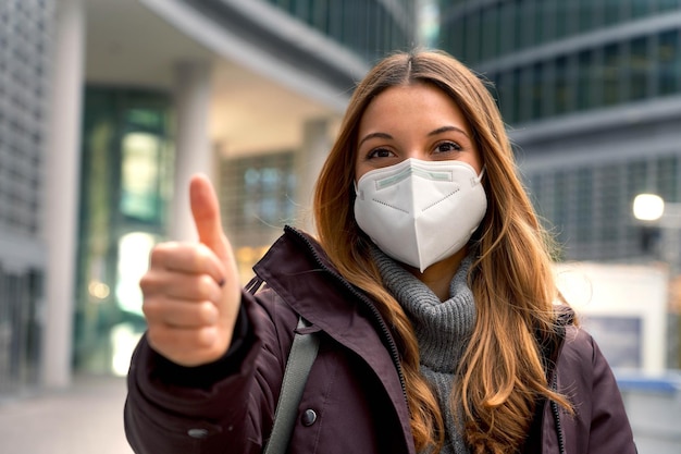 Close-up van een mooie zelfverzekerde vrouw met een beschermend masker KN95 FFP2 die duim omhoog laat zien met moderne stad op de achtergrond en naar de camera kijkt