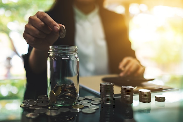 Close-up van een onderneemster die muntstukken in een glaskruik zetten, en geld voor besparing en financieel concept berekenen stapelen