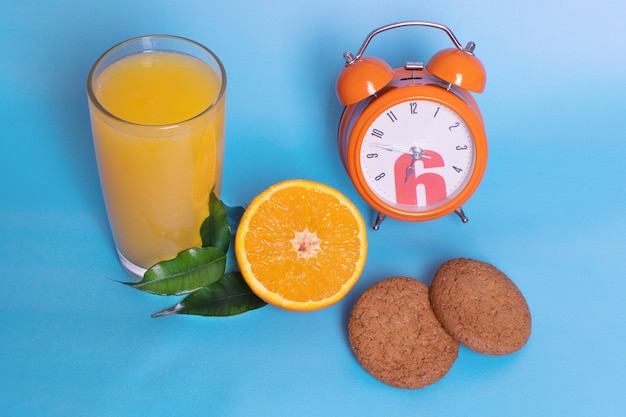 Close-up van een oranje wekker met een glas sinaasappelsap, vers rijp fruit in tweeën gesneden, havermoutkoekjes op een blauwe achtergrond. Biologisch en gezond ontbijtconcept.