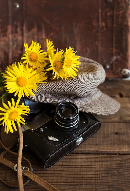 Close-up van een oude camera met een vintage hoed en een boeket gele bloemen op een houten achtergrond.