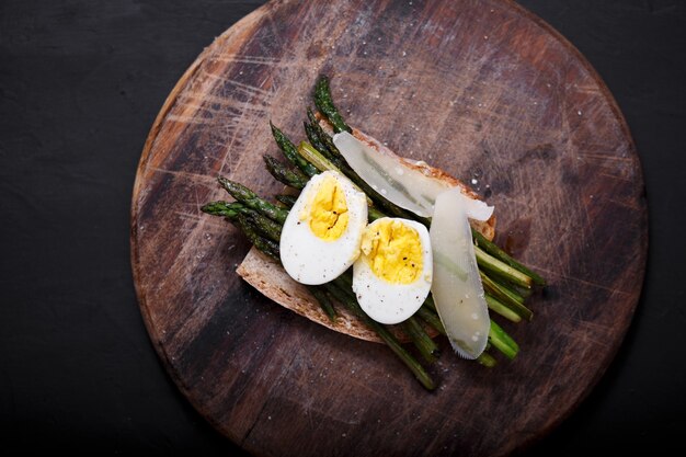 Close up van een plakje met gebakken asperges en gekookt ei in grote houten plaat op stenen achtergrond