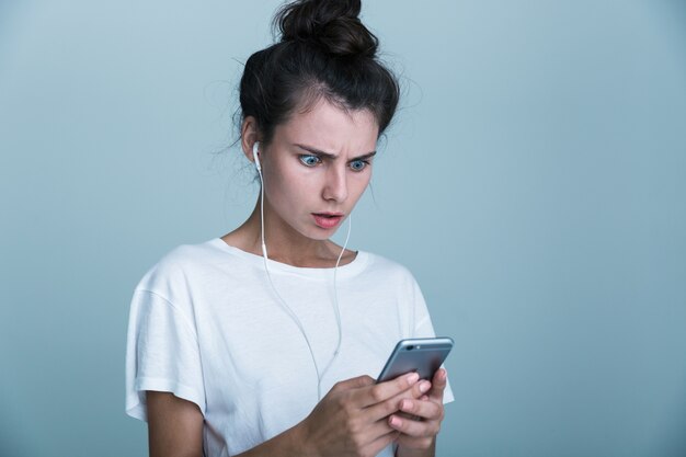 Close-up van een verward casual meisje permanent geïsoleerd op blauwe achtergrond, luisteren naar muziek met koptelefoon en mobiele telefoon kijken