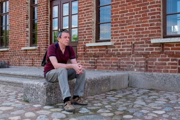 Close-up van een volwassen gepensioneerde in zomerkleren en met een rugzak zit en rust tegen de achtergrond van een rood bakstenen gebouw. Levensstijl. Selectieve aandacht.