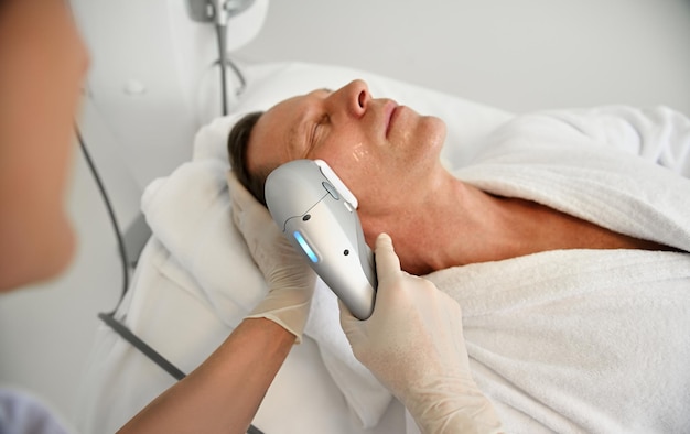 Close-up van een volwassen man die ontspant tijdens schoonheidsprocedures in het wellness-spa-centrum terwijl schoonheidsspecialiste een facelifting anti-aging schoonheidsbehandeling op zijn gezicht doet met behulp van ultrasone medische apparatuur