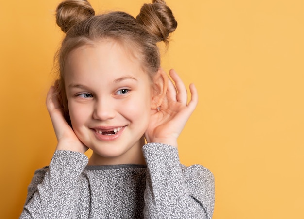 Close-up van een vrolijk lachend meisje raakt haar oren aan en kijkt weg op een gele achtergrond. Concept van plezier en oprechte kinderen emoties. Plaats voor tekst.