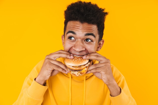 Close-up van een vrolijke jonge afrikaanse tienerjongen die geïsoleerd op geel staat en een grote hamburger eet