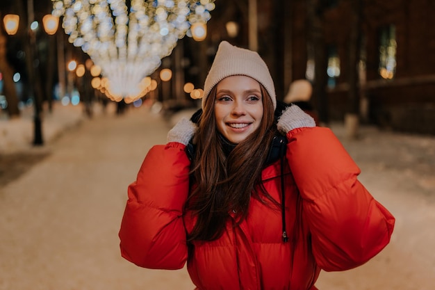 Close-up van een vrolijke jonge vrouw met een hoed en een warm winterjas die op de sneeuwstad poseert