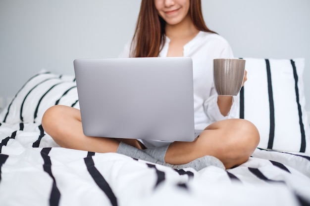 Close-up van een vrouw die en aan laptop computer gebruiken werken, koffie drinken terwijl het zitten op een wit comfortabel bed, het werk van huisconcept