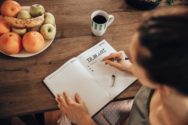 Close-up van een vrouw die in een notitieboekje schrijft en een takenlijst maakt terwijl ze thuis geniet van koffie in de ochtend.