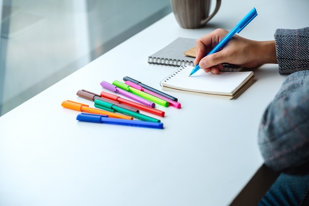Close-up van een vrouw die op een leeg notitieboekje met gekleurde pennen en koffiekop op de lijst schrijft