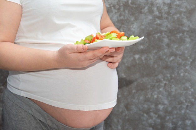 Close-up van een zwangere vrouw die een plaat van gesneden groentesalade op een grijze achtergrond, exemplaarruimte houdt.