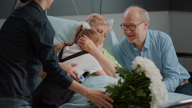 Foto close up van familie verrassende zieke oma in ziekenhuisafdeling bed. vrouw en kind brengen bloemen voor oudere patiënt met ziekte bij bezoek. mensen die gepensioneerde bezoeken. uit de hand geschoten