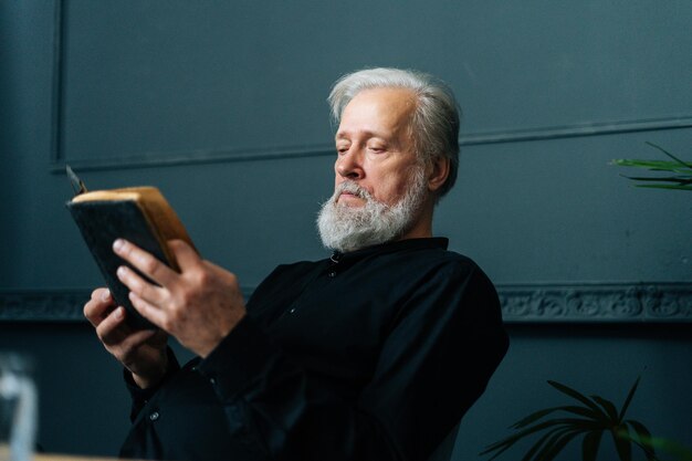 Close-up van gefocuste senior grijsharige oude man die een papieren boek leest dat op een stoel in een donkere woonkamer zit, ontspan en rust met literatuur. Knappe volwassen volwassen man leesboek alleen thuis.