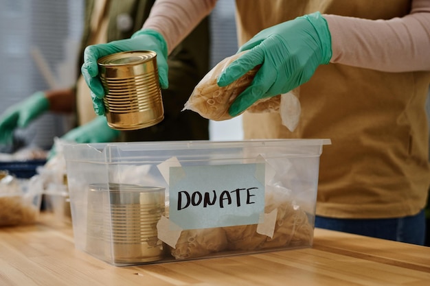 Foto close-up van gehandschoende handen van vrijwilliger die voedselproducten in plastic doos stopt
