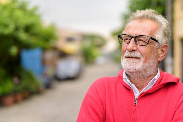 Close up van gelukkig knappe senior bebaarde man glimlachend tijdens het denken en opzoeken met bril buitenshuis