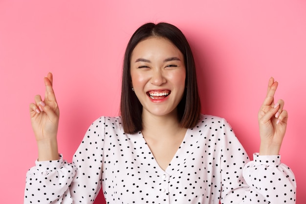 Close-up van gelukkige Aziatische vrouw die een wens doet, vingers gekruist voor geluk houdt en optimistisch glimlacht, die zich over roze bevindt