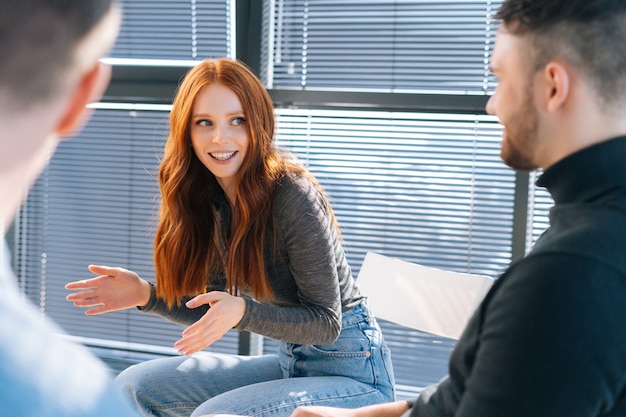 Close-up van gelukkige jonge roodharige zakenvrouw praten en bespreken van nieuwe ideeën met creatieve business team, tijdens brainstormen van start-up projecten in moderne kantoorruimte in de buurt van raam.