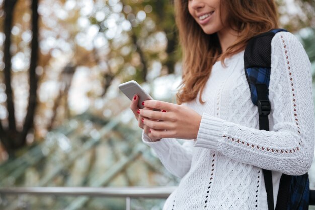 Close-up van gelukkige mooie jonge vrouw met rugzak die mobiele telefoon in park gebruikt