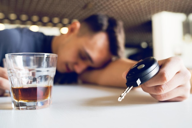 Foto close-up van glas met man die sleutel vasthoudt terwijl hij op tafel slaapt op de achtergrond