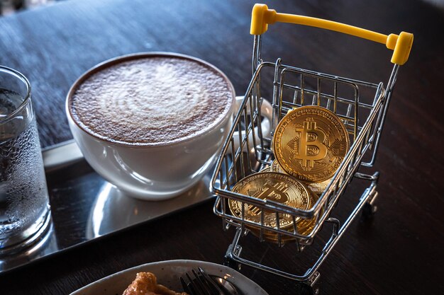 Close-up van gouden bitcoins of cryptocurrency munt of symbolen in winkelwagen en hete koffie latte met latte art melkschuim in de vorm van een zwaan in beker mok op tafel Toekomstig valutaconcept