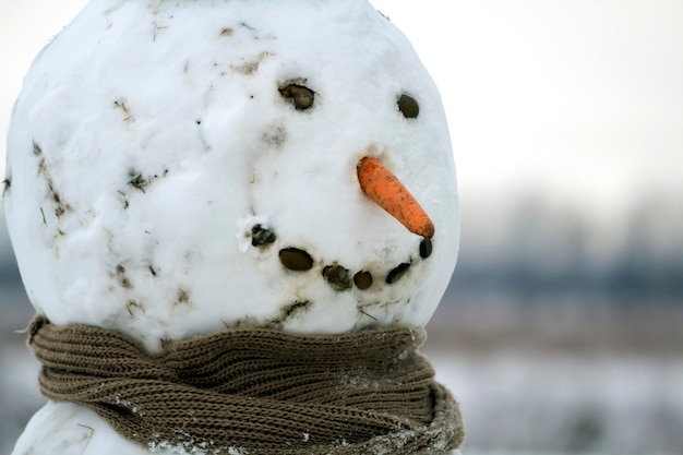 Close-up van groot grappig primitief glimlachend sneeuwmanhoofd met wortelneus, zwarte steenogen en tanden in kindsjaal op licht vage exemplaarruimte in openlucht. Vrolijk kerstfeest en een gelukkig nieuwjaar. A