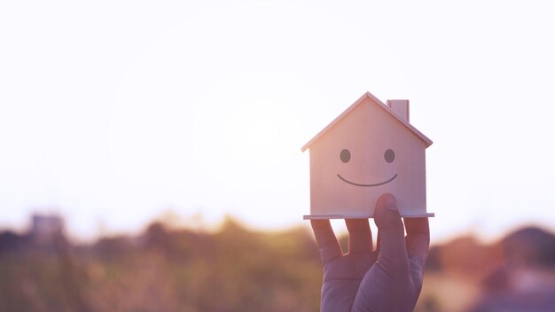 Close-up van hand die houten huis vasthoudt onder zonsondergang met emotie gezicht glimlach