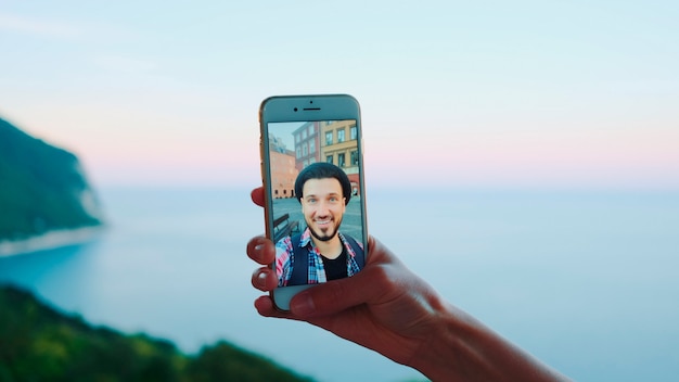 Close-up van hand met smartphone tijdens videogesprek met man voor de zee. Prachtig landschap op de achtergrond.