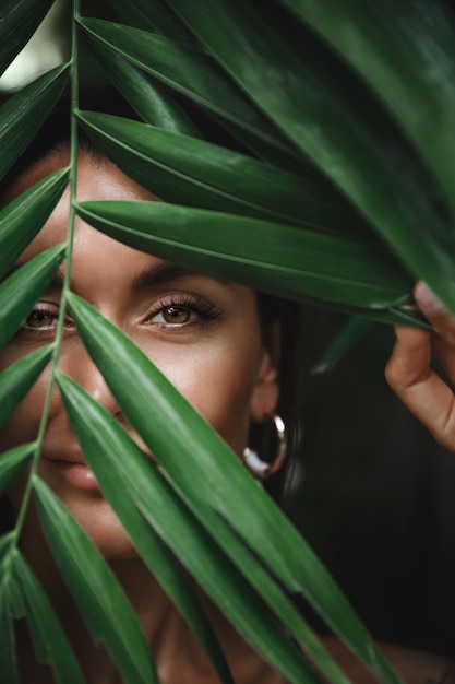 Close-up van het gezicht van de blanke gelooide vrouw zonder make-up, verstopt achter tropische palmbladeren en glimlachen, gluren naar de camera.