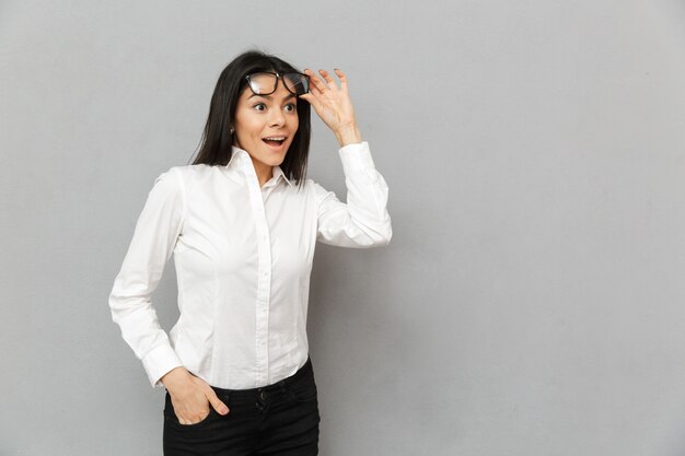 Close-up van het portret van verraste vrouw die bureaukleding draagt die afgezien van onder een bril kijkt, geïsoleerd over grijze achtergrond