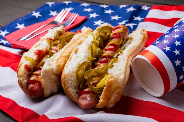 Foto close-up van hotdogs met jalapeno geserveerd op amerikaanse vlag met vork en wegwerpbekers. ongewijzigd, ongezond voedsel, vlees, brood, worst en fastfood.