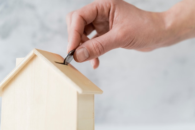 Foto close-up van iemands hand invoegen van de munt in de houten huis spaarpot