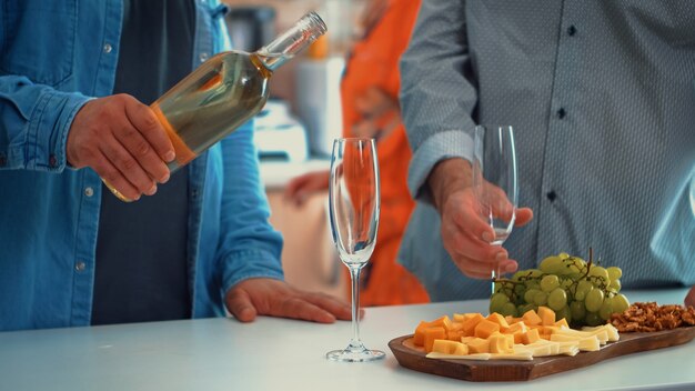 Close up van jonge man gieten witte wijn in glazen. Twee generaties die een kopje champagne proeven in een gezellige eetkamer terwijl vrouwen het gezonde diner bereiden